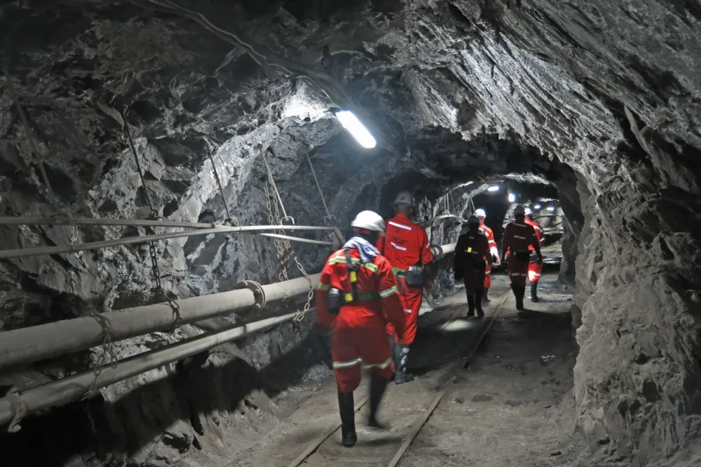 Workers in safety gear inspect a dimly lit underground mine tunnel.