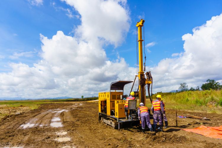 Workers operate a yellow drilling machine on a muddy construction site.