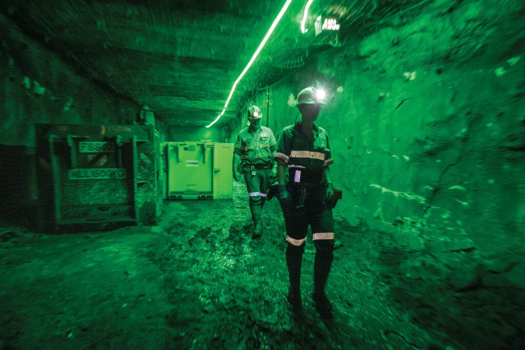 Two miners underground with helmets and safety gear amid green lighting.