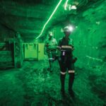 Two miners underground with helmets and safety gear amid green lighting.